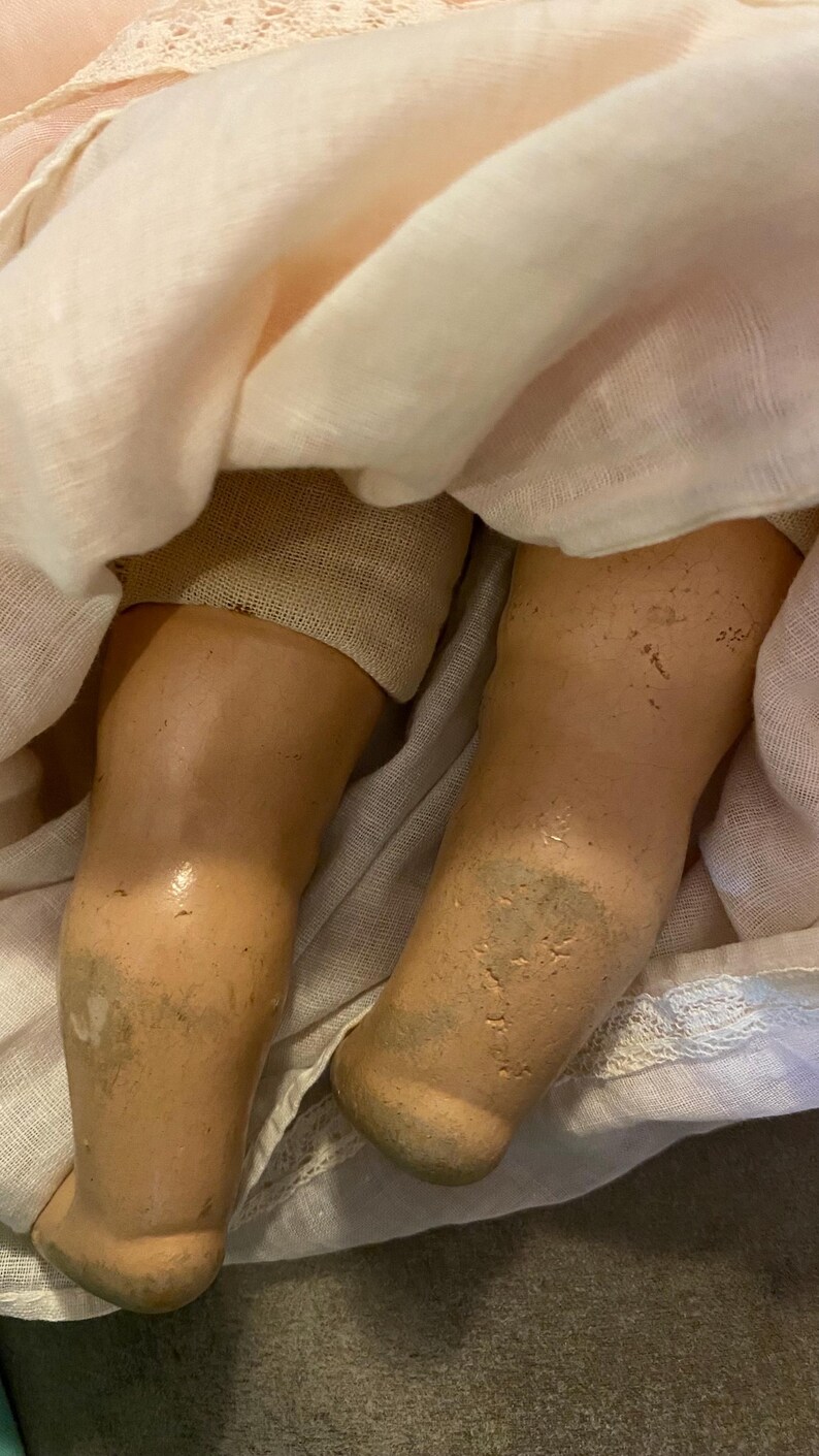 May include: A doll with light tan, textured legs showing signs of wear and discoloration, partially covered by a pale pink fabric garment featuring delicate lace trim at the hem and a soft, layered texture throughout the fabric.