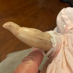 May include: A doll arm with a light beige, cracked composition finish and a visible seam line, attached to a pale pink fabric sleeve featuring delicate white lace trim at the wrist. The arm is positioned horizontally against the fabric body.