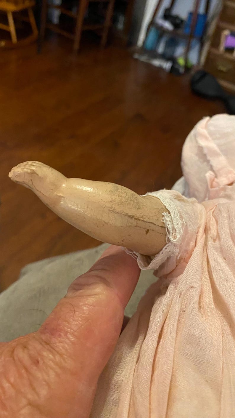 May include: A doll arm with a light beige, cracked composition finish and a visible seam line, attached to a pale pink fabric sleeve featuring delicate white lace trim at the wrist. The arm is positioned horizontally against the fabric body.