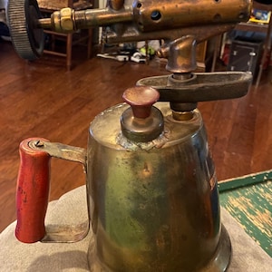 May include: Antique brass blowtorch with a red handle. The torch has a cylindrical fuel tank, a burner head with multiple holes, and a control knob. The handle is made of wood and painted red. The torch is designed for soldering and other heating applications.