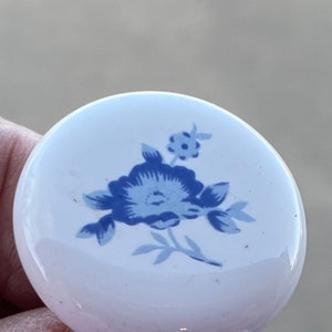May include: A white ceramic knob with a blue floral design. The knob has a smooth, rounded surface and a raised floral pattern.