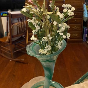 May include: A green glass vase with a swirling design holds a bouquet of white and pink flowers. The vase has a wide mouth and a long, thin neck that curves into a circle at the base.