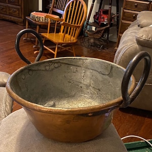 May include: A large copper pot with two black iron handles. The pot has a worn, aged look and is sitting on a brown surface.