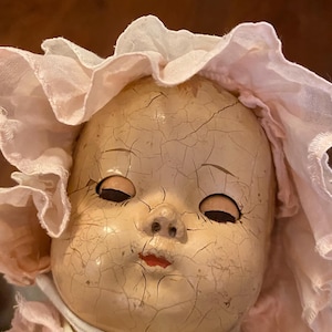 May include: A vintage doll with a heavily crazed, pale face, painted red lips, and dark, closed eyes. The doll wears a light pink ruffled bonnet with a white fabric tie secured under the chin, and a matching pink garment with white lace trim.