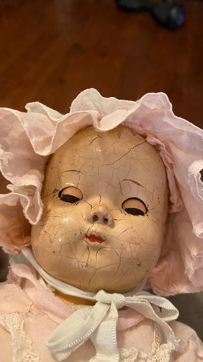 May include: A vintage doll with a heavily crazed, pale face, painted red lips, and dark, closed eyes. The doll wears a light pink ruffled bonnet with a white fabric tie secured under the chin, and a matching pink garment with white lace trim.