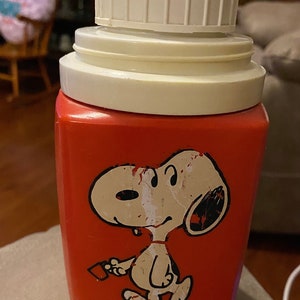 May include: Red and white thermos with a cartoon image of Snoopy holding a red object. The text "Copr. C1958 United Feature Syndicate, Inc." is printed on the bottom of the thermos.