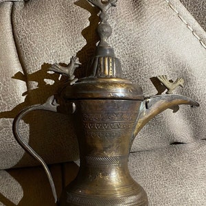 Pode incluir: Uma chaleira de latão antiga com gravuras intrincadas e três alças em forma de pássaro. A chaleira tem um corpo arredondado e uma tampa alta e adornada.
