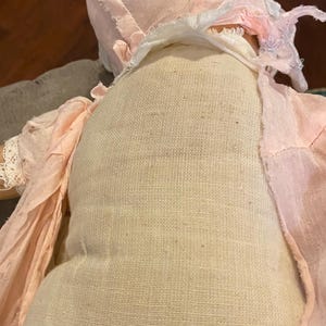 May include: A vintage-style doll featuring a beige fabric torso and a pale pink bonnet with matching fabric accents. The doll is dressed in light pink, textured material with frayed edges and delicate lace trim around the sleeve cuffs.