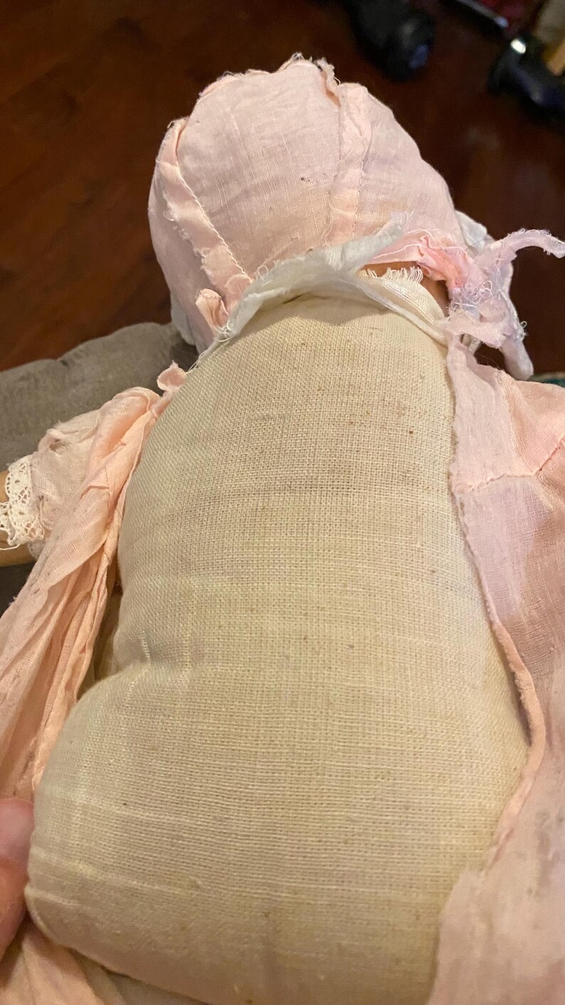 May include: A vintage-style doll featuring a beige fabric torso and a pale pink bonnet with matching fabric accents. The doll is dressed in light pink, textured material with frayed edges and delicate lace trim around the sleeve cuffs.