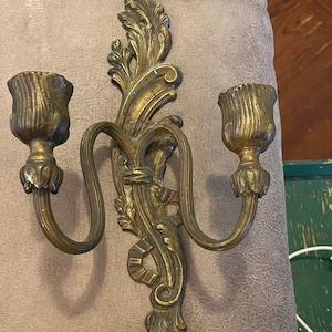 May include: A gold-toned metal wall sconce with two candle holders. The sconce has a decorative, ornate design with a floral motif.