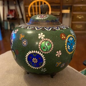 May include: A green cloisonné jar with a lid, decorated with colorful floral and geometric patterns. The jar has three small feet and is made of metal.
