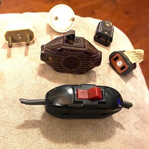 May include: A collection of vintage electrical plugs and switches. Includes a brown bakelite switch with the text "CAT. NO. 750", a black inline switch with a red button, and various plug types in cream and black.