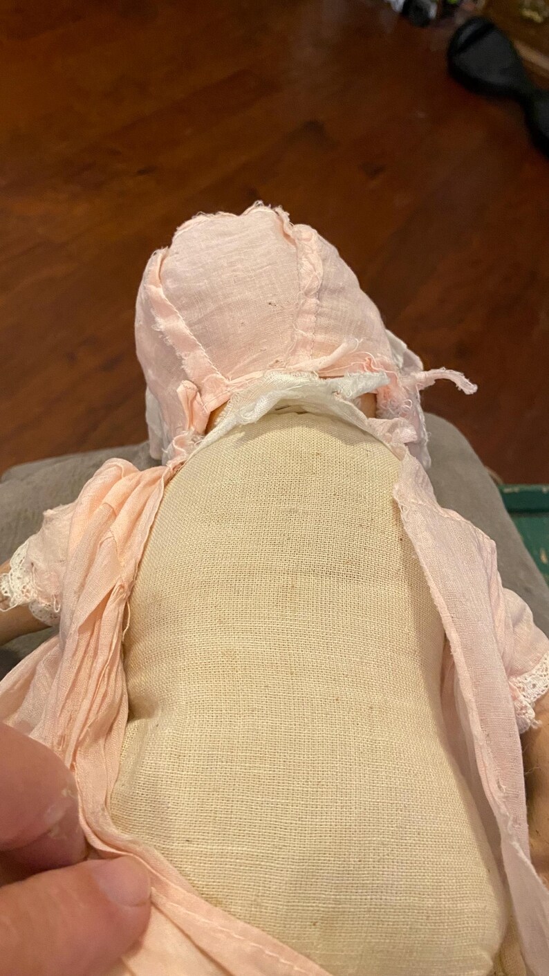 May include: A doll with a beige fabric torso wearing a pale pink bonnet and a matching pale pink dress with delicate lace trim on the sleeves. The doll is viewed from the back, showing the texture of the woven fabric and the layered pink garment.