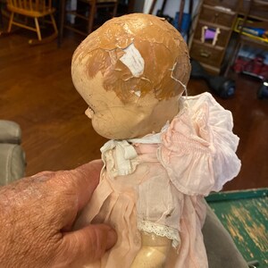 May include: A vintage doll with a cracked, peeling tan painted head and a light pink fabric dress featuring a ruffled collar, short sleeves with lace trim, and a white ribbon tie at the neck. The doll has a smooth, light-colored arm visible.