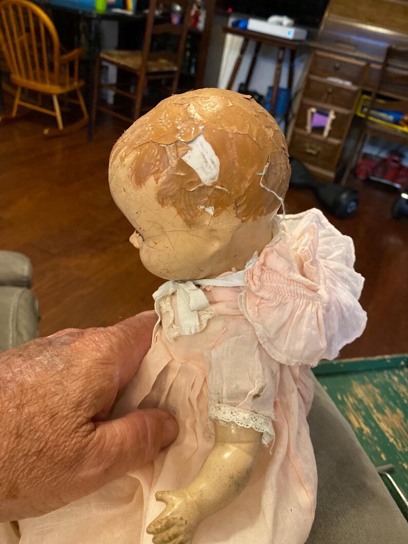 May include: A vintage doll with a cracked, peeling tan painted head and a light pink fabric dress featuring a ruffled collar, short sleeves with lace trim, and a white ribbon tie at the neck. The doll has a smooth, light-colored arm visible.