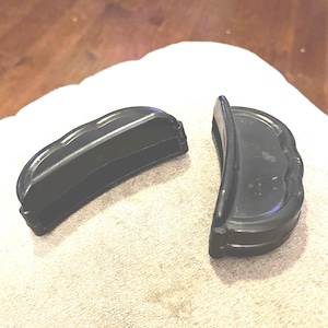 May include: Two black, semi-circular plastic components with a scalloped edge design. Each piece features a hollow, rectangular opening along the inner side and a smooth, curved exterior surface with a tiered base structure.