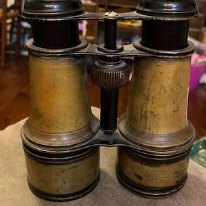 May include: Antique brass and black binoculars. The binoculars have a central focusing mechanism and are made of brass and black metal. The brass has a worn, aged appearance. The binoculars are resting on a tan surface.