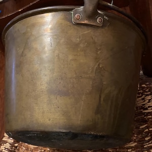 May include: A vintage brass bucket with a handle. The bucket has a rounded shape and a dark, aged patina. The handle is attached with rivets. The bucket is likely used for decorative purposes or as a planter.