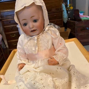 May include: A porcelain baby doll wearing a white dress with lace trim and a pink sash. The doll has blue eyes and a painted face.