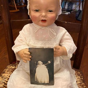 May include: Vintage porcelain baby doll wearing a white dress and bonnet, seated on a wooden chair. The doll holds a black and white photograph of a baby. The doll has rosy cheeks and a slightly open mouth.