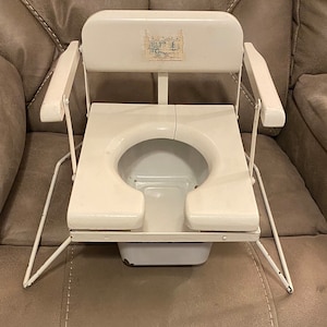 May include: White potty chair with a white metal frame and a white plastic seat. The chair has a back rest and arm rests. The chair has a label that says "Little Toidey".