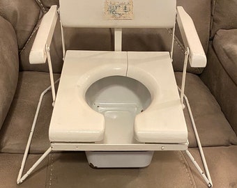 May include: White potty chair with a white metal frame and a white plastic seat. The chair has a back rest and arm rests. The chair has a label that says "Little Toidey".