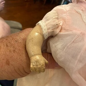 May include: A vintage doll with a pale, weathered composition arm showing signs of age and wear. The doll is dressed in a light pink fabric garment featuring delicate white lace trim along the sleeve and a matching pink bonnet on its head.