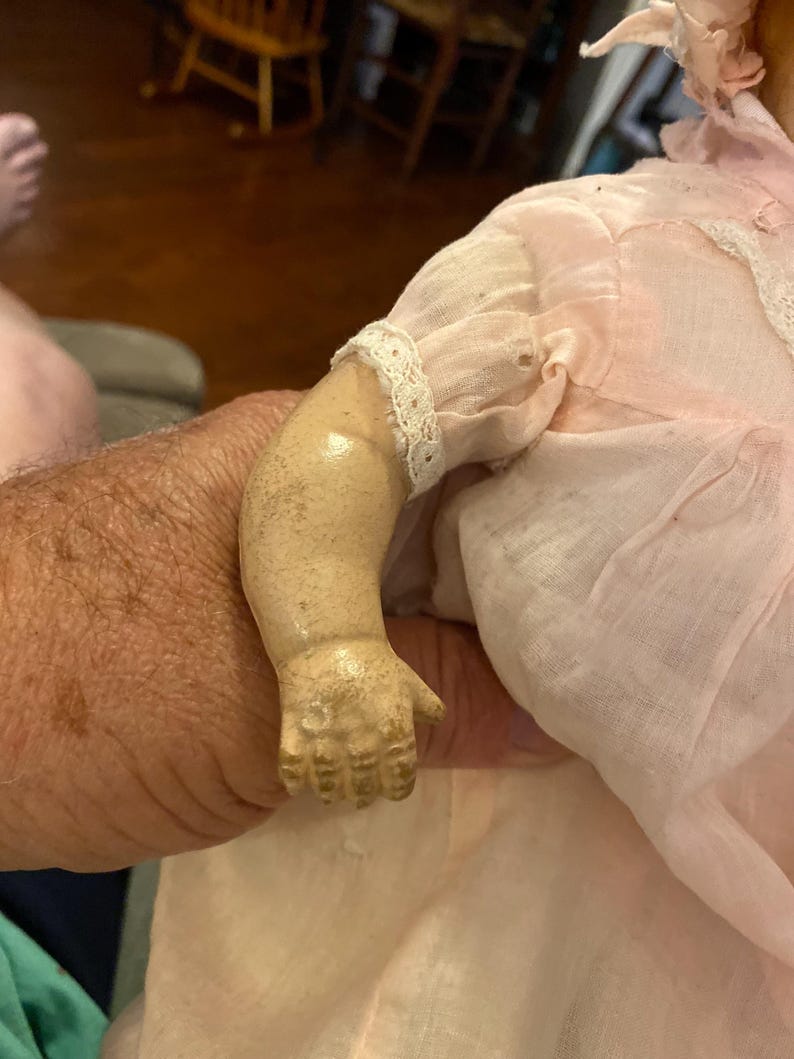 May include: A vintage doll with a pale, weathered composition arm showing signs of age and wear. The doll is dressed in a light pink fabric garment featuring delicate white lace trim along the sleeve and a matching pink bonnet on its head.