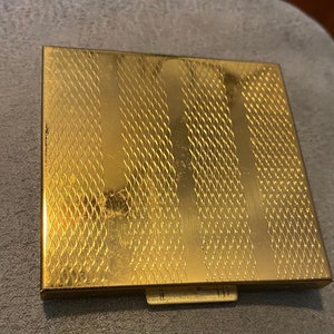 May include: A square gold-tone metal compact case featuring a textured surface with alternating vertical bands of smooth polished metal and a fine, wavy cross-hatched pattern. The case includes a small rectangular latch on the front edge.