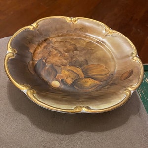 May include: A decorative ceramic plate with a gold rim and a painted design of nuts and fruit. The plate is signed "Art Daniel".