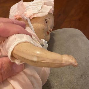 May include: A vintage doll with a cracked, aged face and painted features, wearing a pale pink fabric bonnet with ruffled edges and a matching pale pink outfit. The doll has a light-toned, smooth arm and is dressed in simple, light-colored clothing.