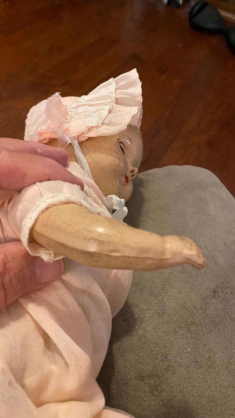 May include: A vintage doll with a cracked, aged face and painted features, wearing a pale pink fabric bonnet with ruffled edges and a matching pale pink outfit. The doll has a light-toned, smooth arm and is dressed in simple, light-colored clothing.