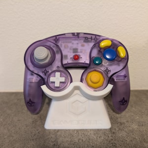 Könnte beinhalten: Ein transparent-violetter Videospiel-Controller mit bunten Knöpfen, der auf einem weißen Ständer steht. Der Controller hat einen grauen Joystick, ein weißes kreuzförmiges Steuerkreuz und gelbe, blaue und grüne Knöpfe. Der Ständer trägt den Text "GAMECUBE".
