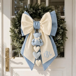 Può includere: Una ghirlanda decorativa con un grande fiocco a strati in tessuto color crema e azzurro. Un orsetto di peluche azzurro pende dal centro, seguito da tre cuori decorativi pendenti. Il pannello in tessuto color crema presenta le lettere ricamate EBL.