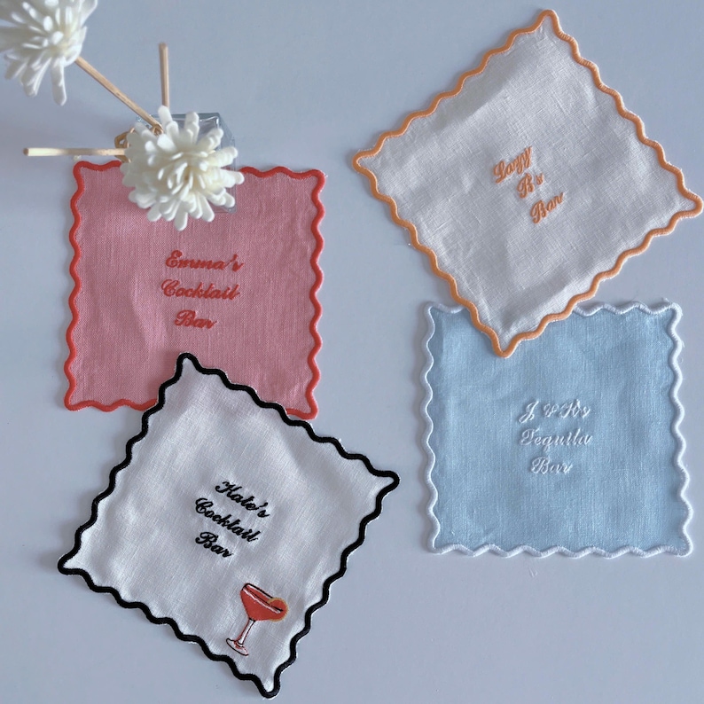 May include: Four square cocktail napkins in pink, white, and blue with scalloped edges. Each napkin has embroidered text: "Emma's Cocktail Bar", "Lazy R's Bar", "Hale's Cocktail Bar", and "J & R's Tequila Bar".