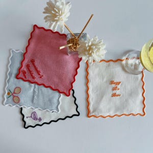 May include: A collection of embroidered square coasters with scalloped edges. The coasters are in various colors, including pink, white, and light blue. Some coasters have text that reads "Danielle's Cocktail Bar" and "Lazy B's Bar". Other coasters have embroidered tennis rackets and a flamingo.
