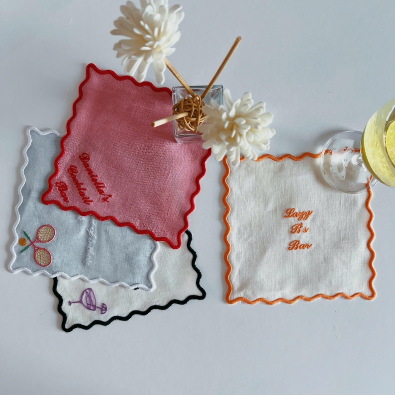 May include: A collection of embroidered square coasters with scalloped edges. The coasters are in various colors, including pink, white, and light blue. Some coasters have text that reads "Danielle's Cocktail Bar" and "Lazy B's Bar". Other coasters have embroidered tennis rackets and a flamingo.