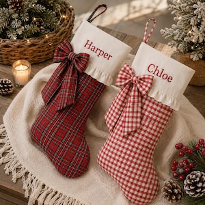 Op de afbeelding: Twee kerstsokken met witte boorden waarop de namen Harper en Chloe zijn geborduurd. De linker sok heeft een rood ruitpatroon met een bijpassende strik, terwijl de rechter sok een rood-wit gingham patroon heeft met een bijpassende strik.