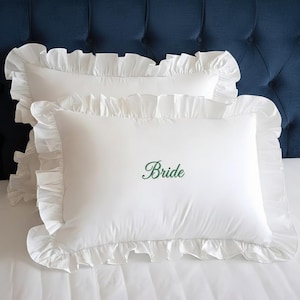 Puede incluir: Dos fundas de almohada rectangulares blancas con bordes con volantes. La palabra "Bride" está bordada en verde en la parte delantera de una almohada. Las almohadas están sobre una cama blanca con un cabecero azul en el fondo.