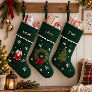 Könnte beinhalten: Drei dunkelgrüne Weihnachtsstrümpfe aus Samt hängen an einer Holzleiste. Jeder Strumpf hat einen weißen, gewellten Umschlag mit den eingestickten Namen Lucas, Mom und Dad. Sie sind mit weißen Schneeflocken und Stickmotiven verziert.