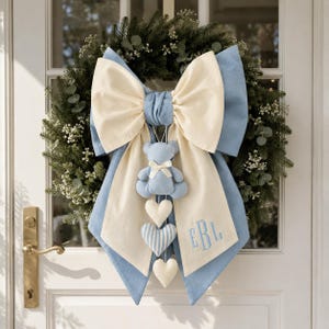 Peut inclure: Une couronne décorative avec un grand nœud superposé en tissu crème et bleu clair. Un ours en peluche bleu clair est suspendu au centre, suivi de trois cœurs décoratifs. Le panneau en tissu crème porte les lettres brodées EBL.