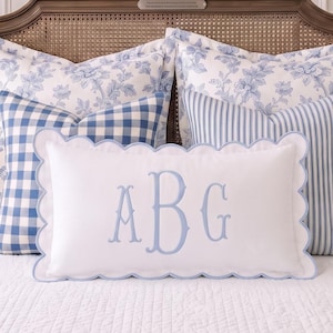 May include: A rectangular white throw pillow with a scalloped edge and light blue piping. The center features a large, embroidered light blue monogram with the letters ABG in a decorative serif font. The pillow is set against other pillows.