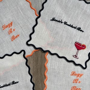May include: White linen cocktail napkins with black and orange scalloped edges. One napkin features a red cocktail graphic, while others have embroidered text that reads "Lazy B's Bar" and "Janie's Cocktail Bar".