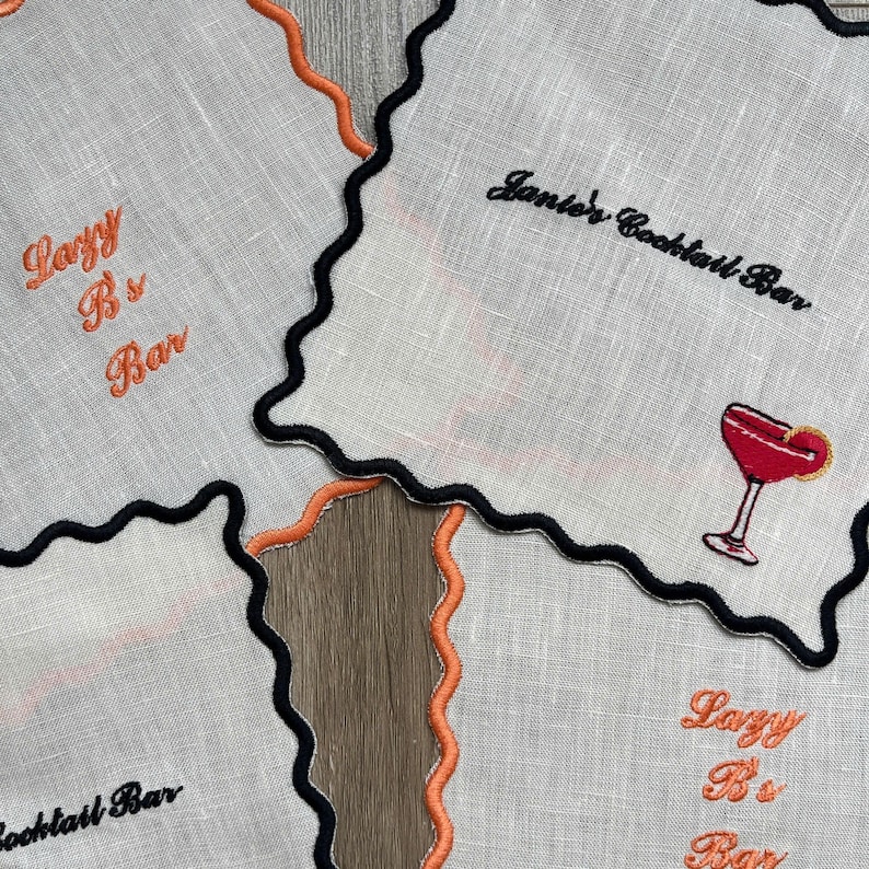 May include: White linen cocktail napkins with black and orange scalloped edges. One napkin features a red cocktail graphic, while others have embroidered text that reads "Lazy B's Bar" and "Janie's Cocktail Bar".