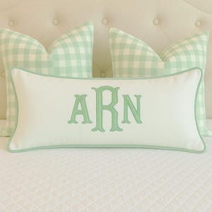 May include: A white rectangular cushion with a pale green border and embroidered initials "ARN" in a matching pale green font. Two gingham-patterned cushions in pale green and white are behind it. The cushions are on a white quilted surface.