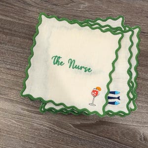 May include: White handkerchiefs with green scalloped edges. The text "The Nurse" is embroidered in green. A cocktail glass and three blue fish are also embroidered on the handkerchief.
