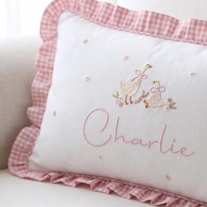 May include: White rectangular cushion with a pink gingham frill border. Embroidered with two geese wearing pink bows and the name "Charlie" in pink script. Small embroidered flowers are scattered across the cushion.