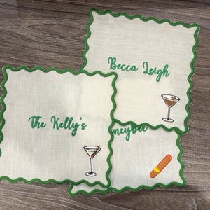 May include: Four white linen cocktail napkins with green scalloped edges. Each napkin has embroidered text and a cocktail or cigar illustration. The text includes "The Kelly's", "Becca Leigh", and "Honeybee".