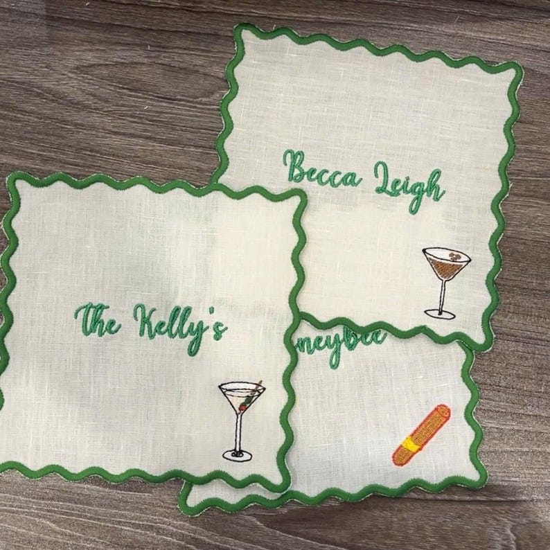 May include: Four white linen cocktail napkins with green scalloped edges. Each napkin has embroidered text and a cocktail or cigar illustration. The text includes "The Kelly's", "Becca Leigh", and "Honeybee".