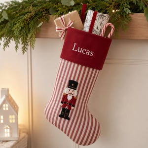 Op de afbeelding: Een kerstkous met een rood-wit gestreept patroon en een effen rode fluwelen boord, geborduurd met de naam Lucas in wit garen. Een applicatie van een notenkrakerfiguur is in het midden aan de voorzijde geplaatst, met cadeaus en een zuurstok.
